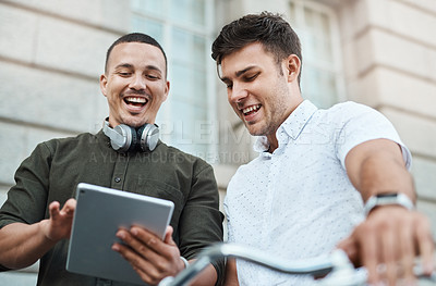 Buy stock photo Shot of two young businessmen using a digital tablet together while riding a bicycle in the city