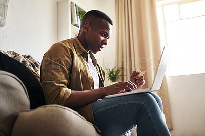 Buy stock photo Cropped shot of a handsome young man using a laptop and a credit card to shop online while sitting on his couch at home