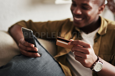 Buy stock photo Cropped shot of a handsome young man smiling while using a smartphone and a credit card to shop online in his living room at home