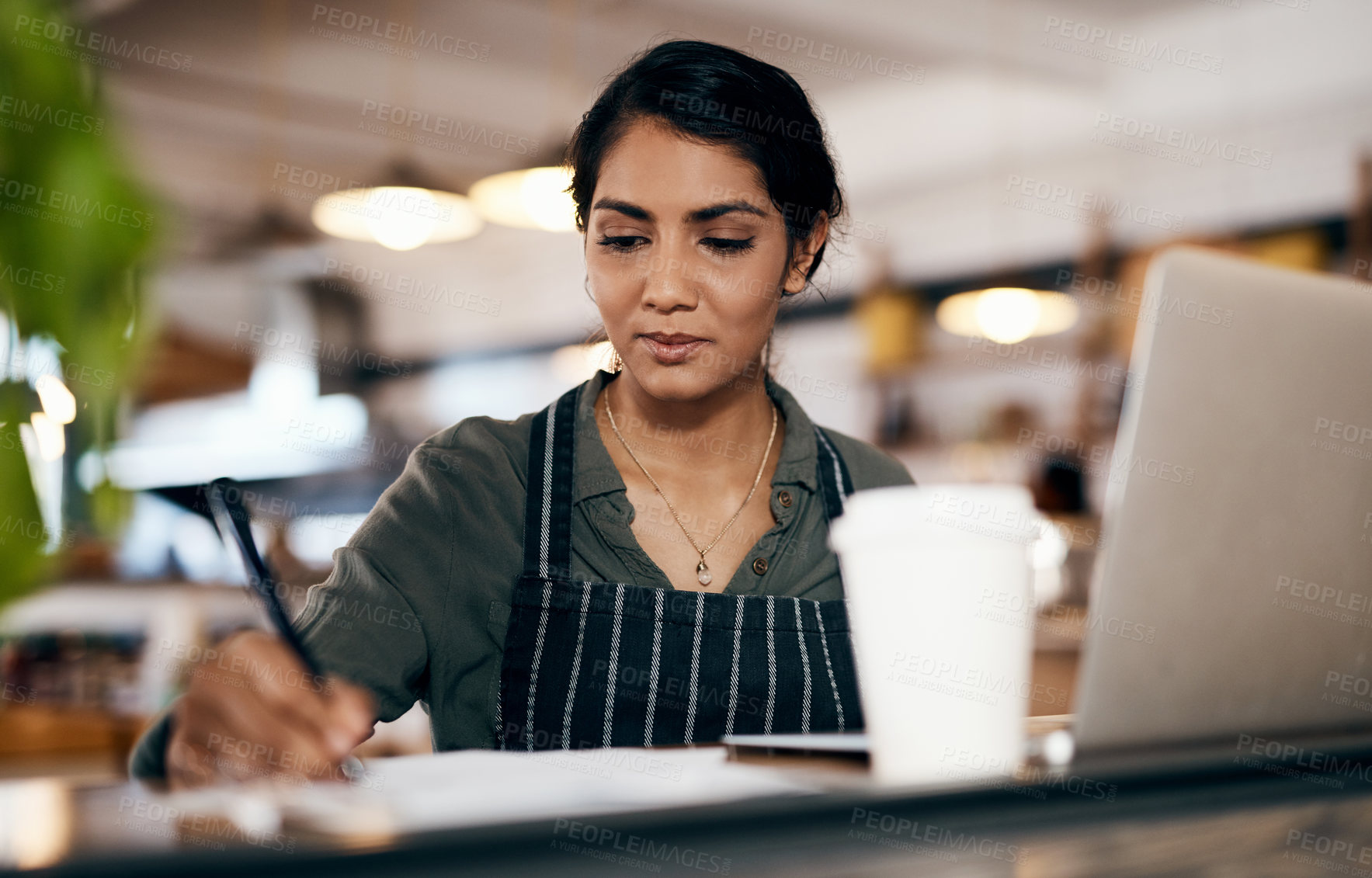 Buy stock photo Small business owner working on a budget in her coffee shop and writing a list of stock to order. Serious female entrepreneur developing a growth strategy for her cafe startup