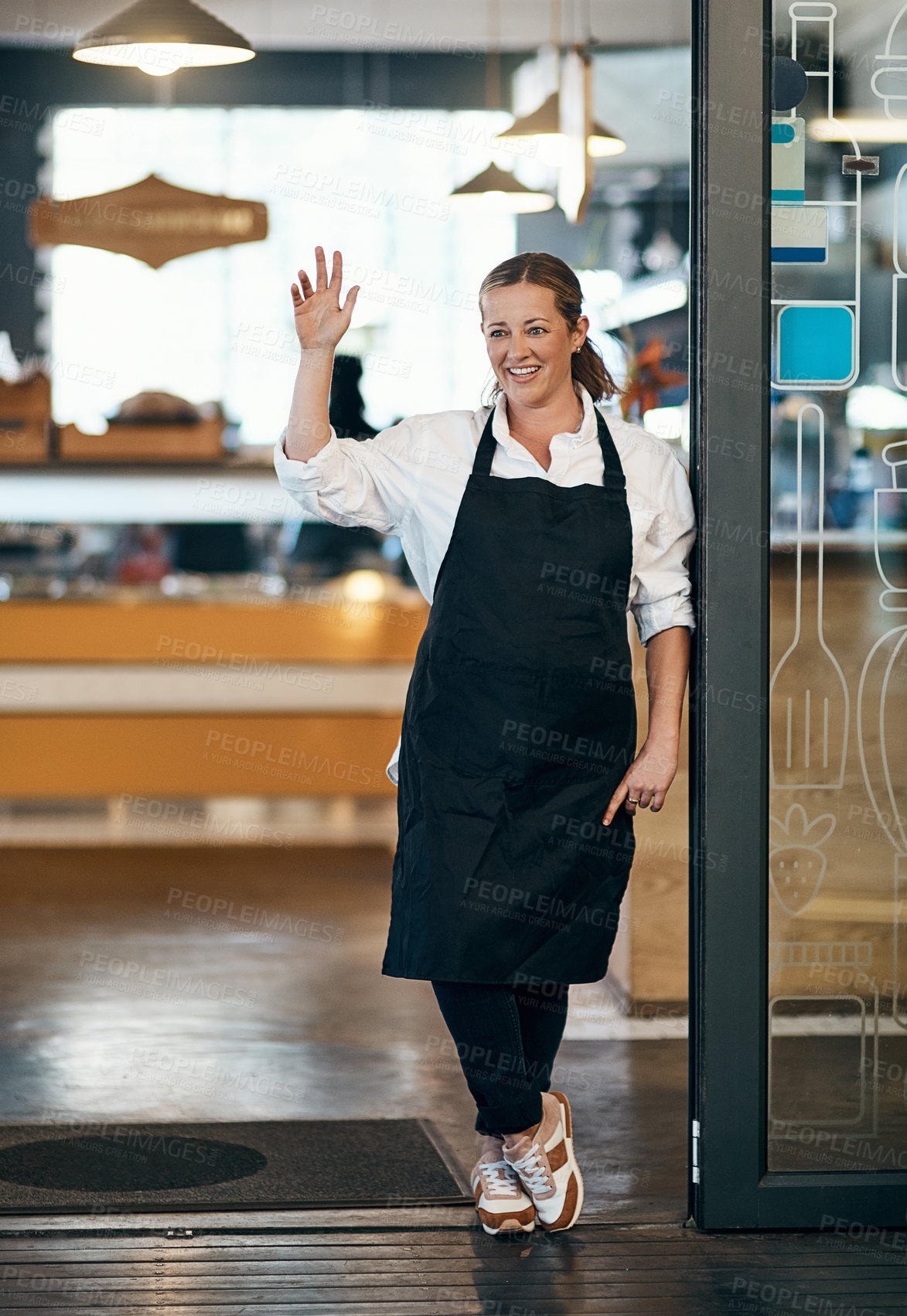 Buy stock photo Shot of a mature woman standing at the entrance of her cafe and waving