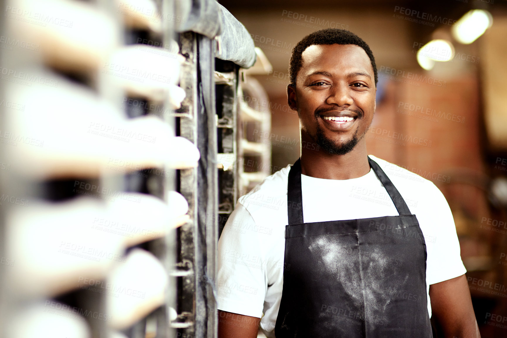 Buy stock photo Black man, portrait and bakery with apron for production, fresh bread or pastry by trolley at shop. Young African, male person or professional baker with smile for small business or startup at store