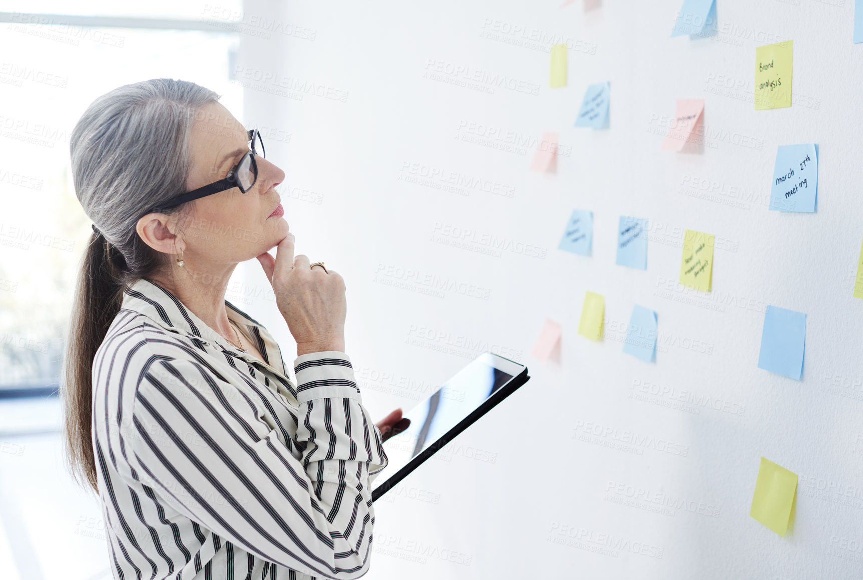 Buy stock photo Shot of a mature businesswoman using a digital tablet while brainstorming with notes on a wall in an office