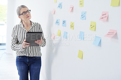 Buy stock photo Shot of a mature businesswoman using a digital tablet while brainstorming with notes on a wall in an office