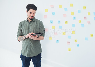 Buy stock photo Shot of a young businessman using a digital tablet while standing against a wall with notes in an office