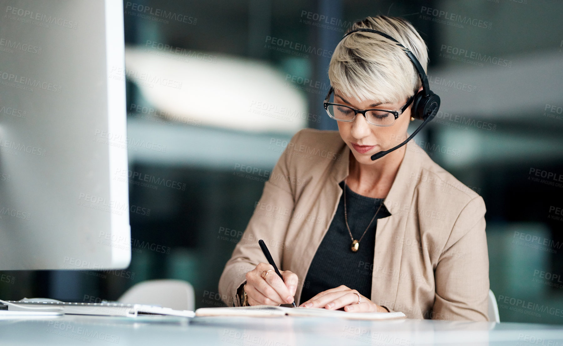 Buy stock photo Shot of a young businesswoman wearing a headset while writing notes in an office