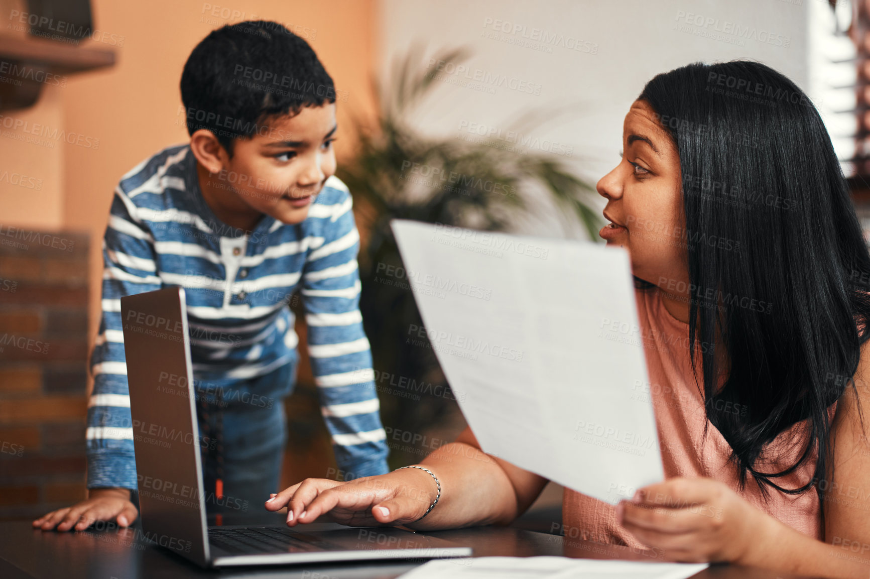 Buy stock photo Shot of a mother using a laptop while going through paperwork with her son alongside her at home