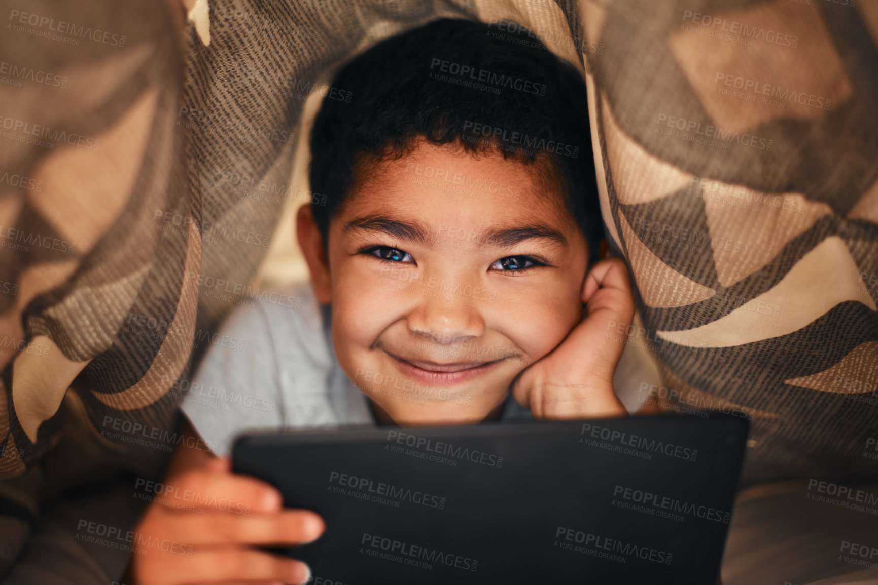 Buy stock photo Portrait of an adorable little boy using a digital tablet while lying under a blanket at home