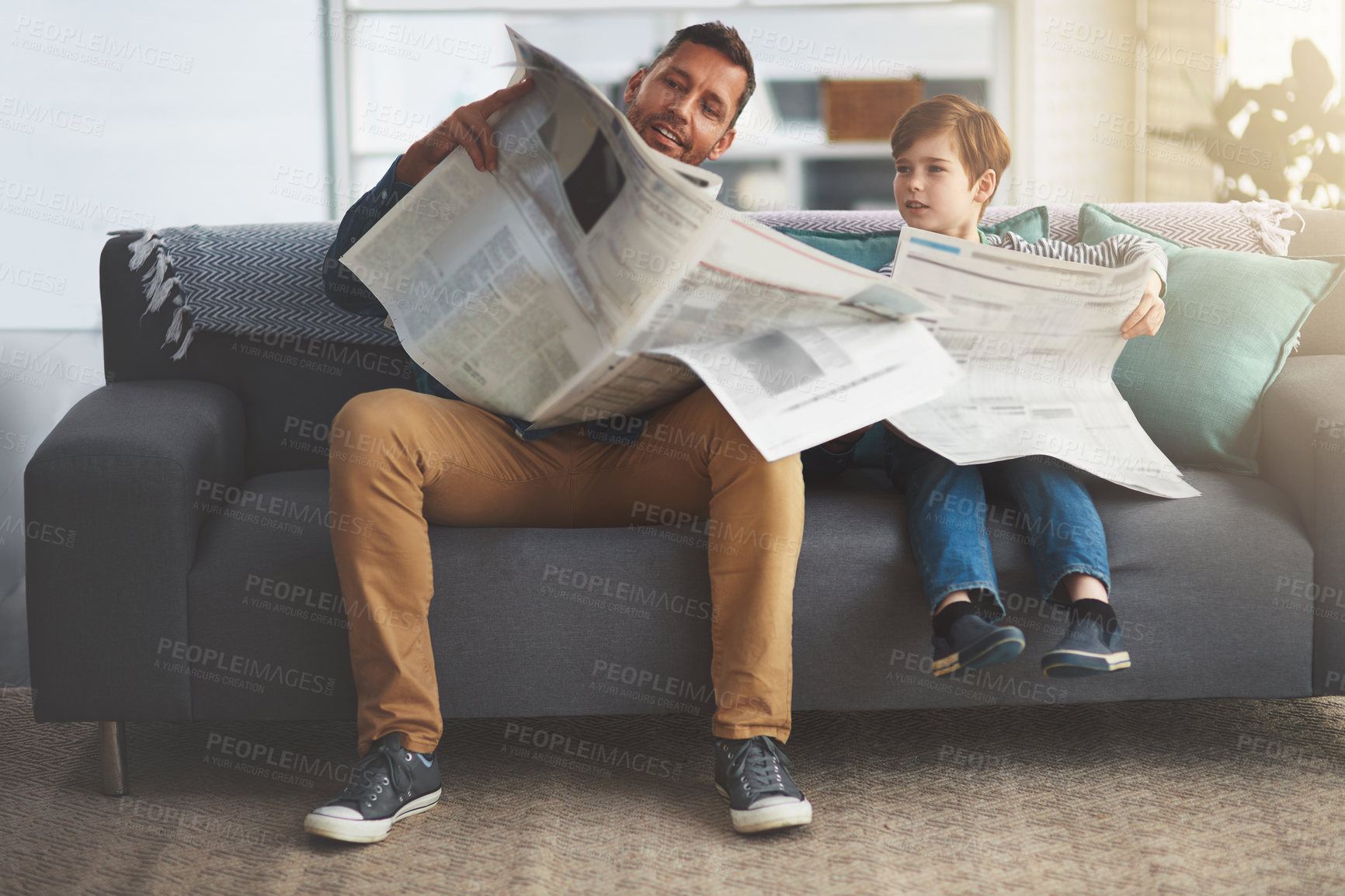 Buy stock photo Shot of a carefree little boy and his father reading the newspaper while being seated on the sofa at home during the day