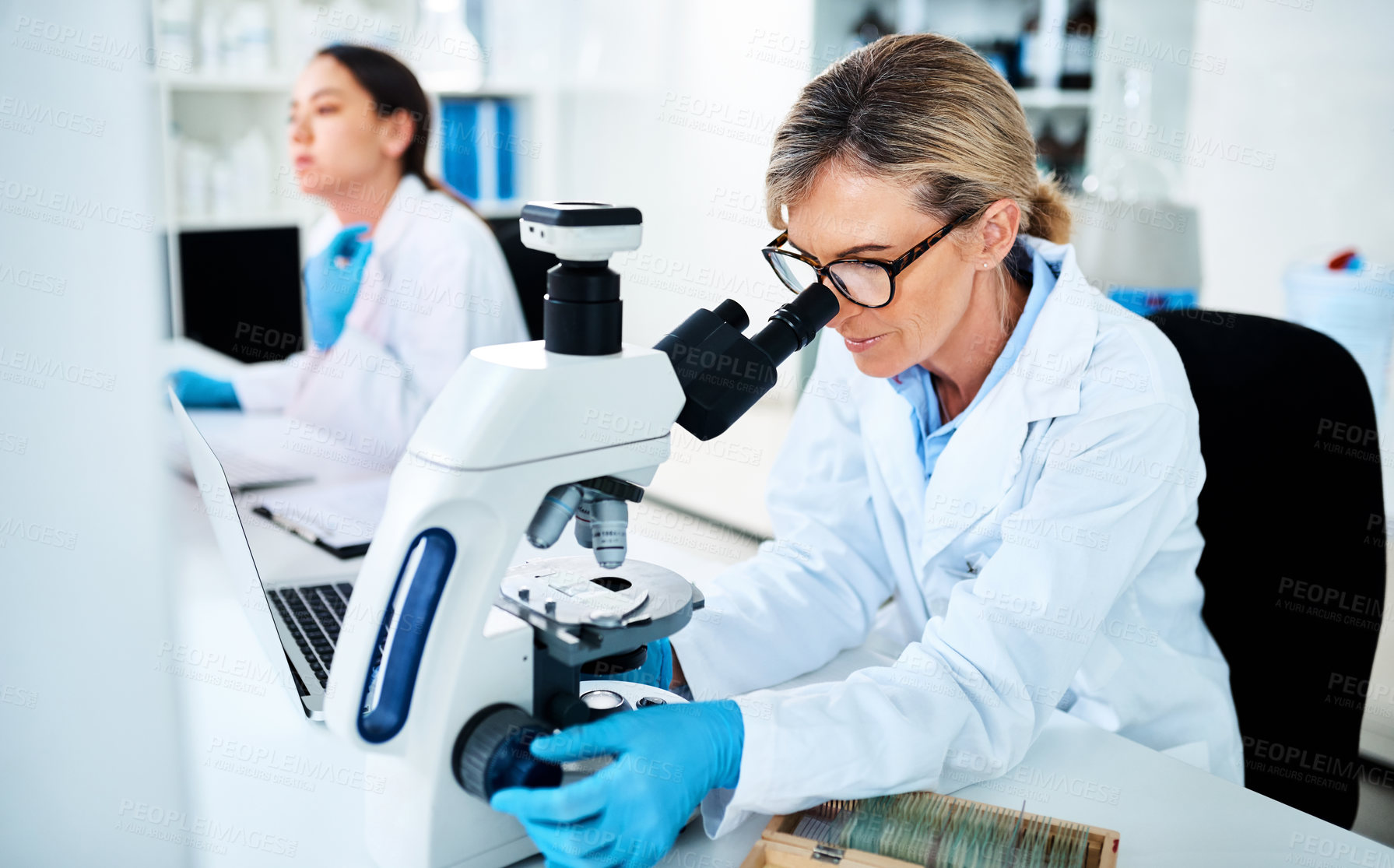 Buy stock photo Shot of a mature scientist using a microscope in a lab with her colleague in the background