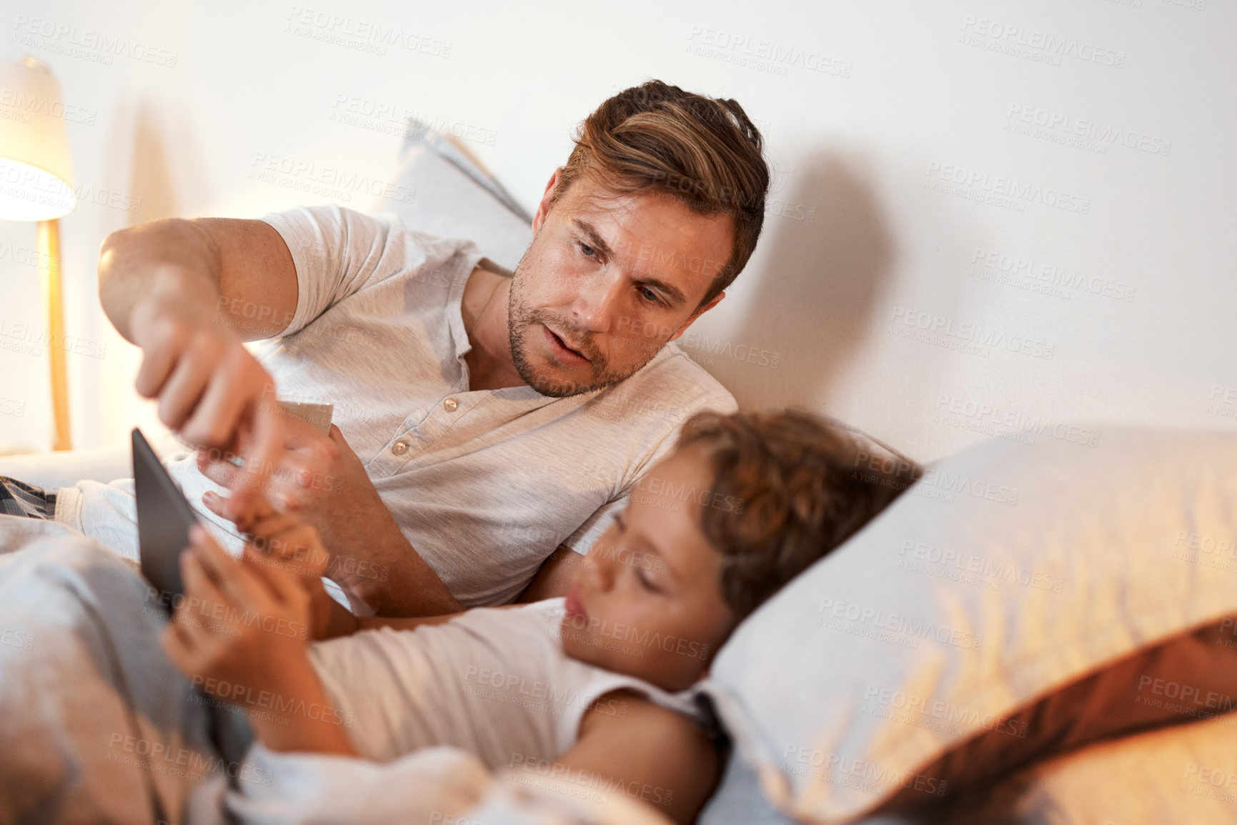 Buy stock photo Shot of a young boy using a digital tablet while lying in bed with his father