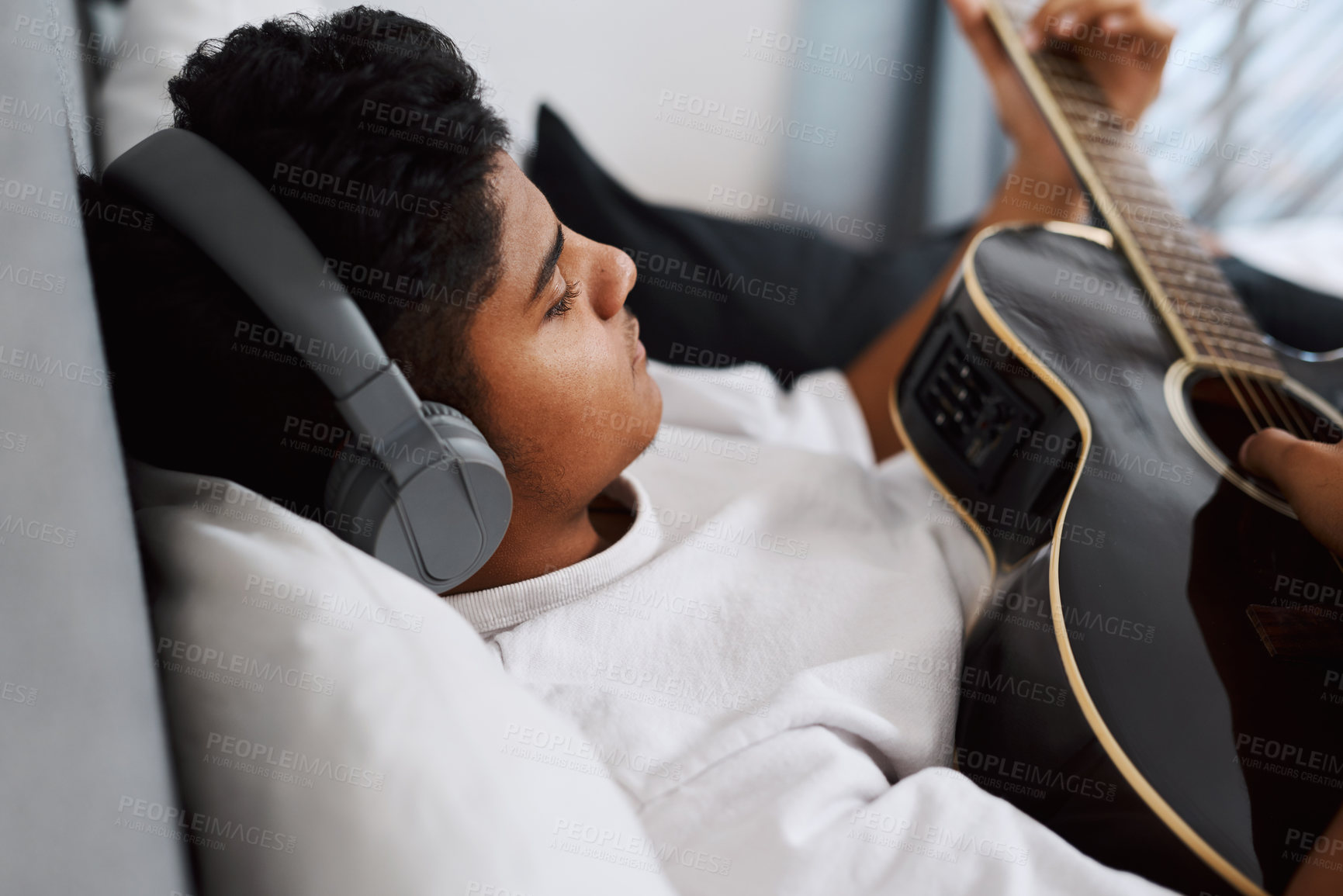 Buy stock photo Shot of a young man wearing headphones while playing the guitar at home