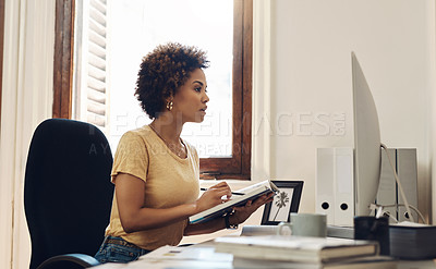 Buy stock photo Shot of a young businesswoman working on a computer while going through notes from a book in an office