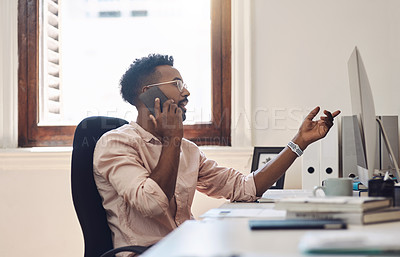 Buy stock photo Shot of a young businessman talking on a cellphone while working on a computer in an office