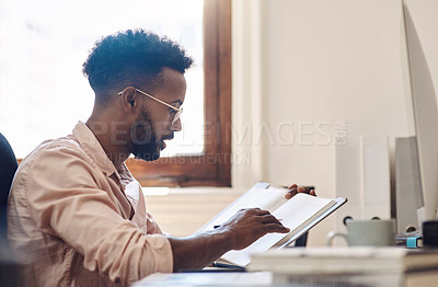 Buy stock photo Shot of a young businessman going through notes from a book while working in an office