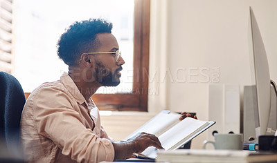 Buy stock photo Shot of a young businessman working on a computer while going through notes from a book in an office