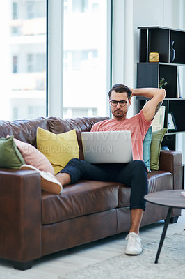 Buy stock photo Shot of a young man using a laptop while relaxing on a sofa
