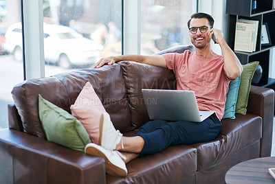 Buy stock photo Shot of a young man using a laptop and smartphone while relaxing on a sofa