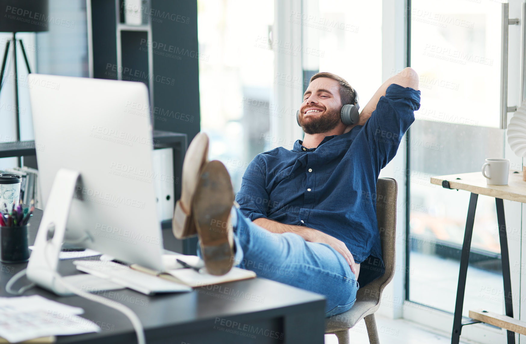 Buy stock photo Shot of a happy young businessman using headphones while relaxing at her desk in a modern office