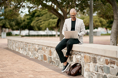 Buy stock photo Shot of a mature businessman sitting on a wall and using a laptop against a city background