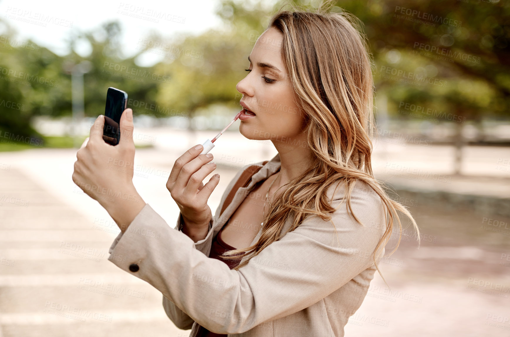 Buy stock photo Shot of a young businesswoman applying lipstick using a smartphone against a city background