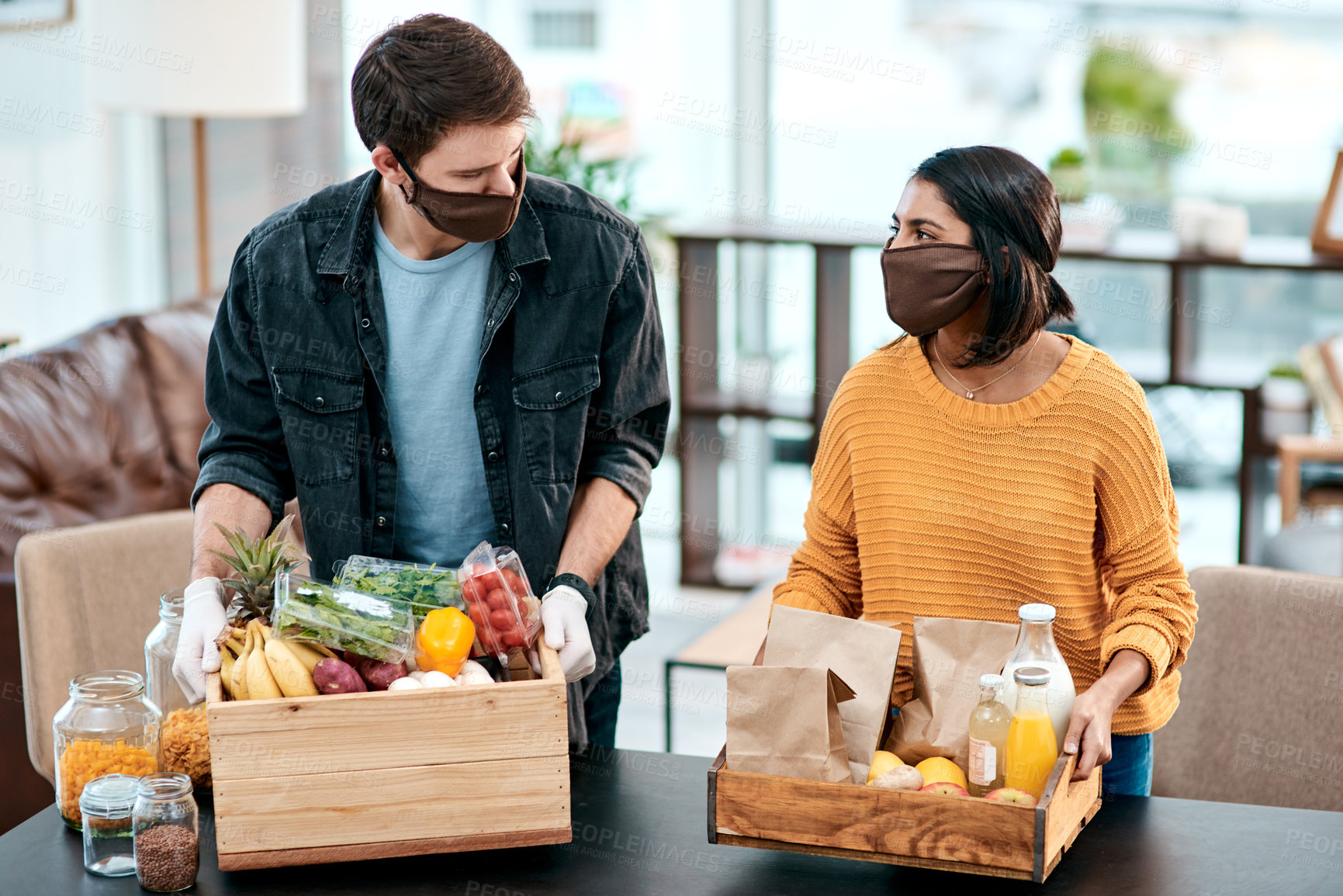 Buy stock photo Shot of a young couple wearing masks while unpacking their groceries at home