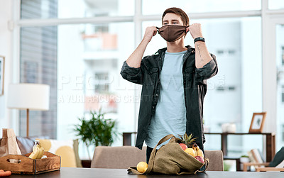 Buy stock photo Shot of a young man putting on a mask before unpacking his groceries at home