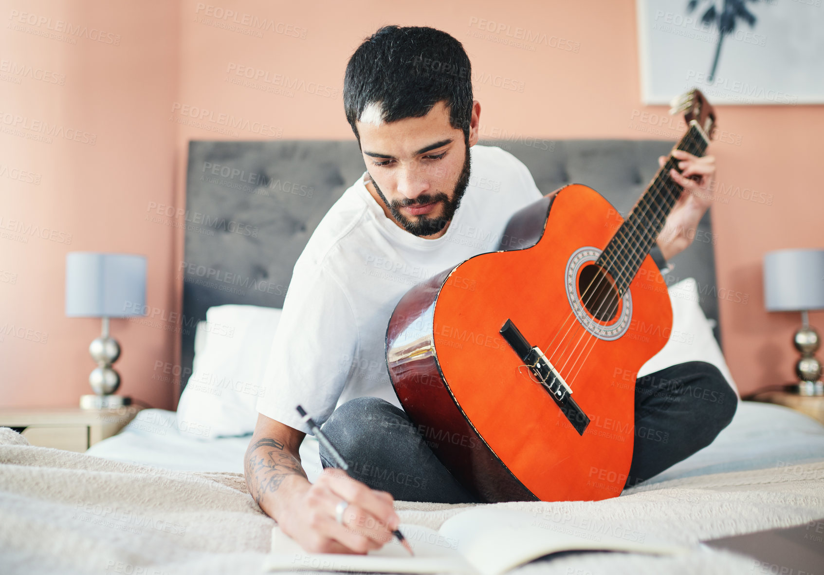 Buy stock photo Shot of a man writing in his notebook while playing the guitar at home