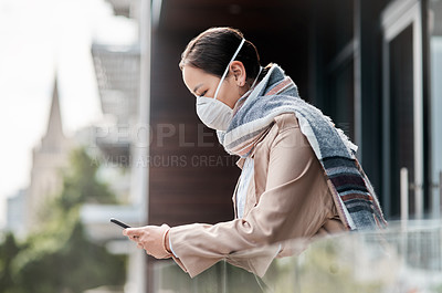 Buy stock photo Shot of a young woman wearing a mask and using a smartphone on the balcony at home