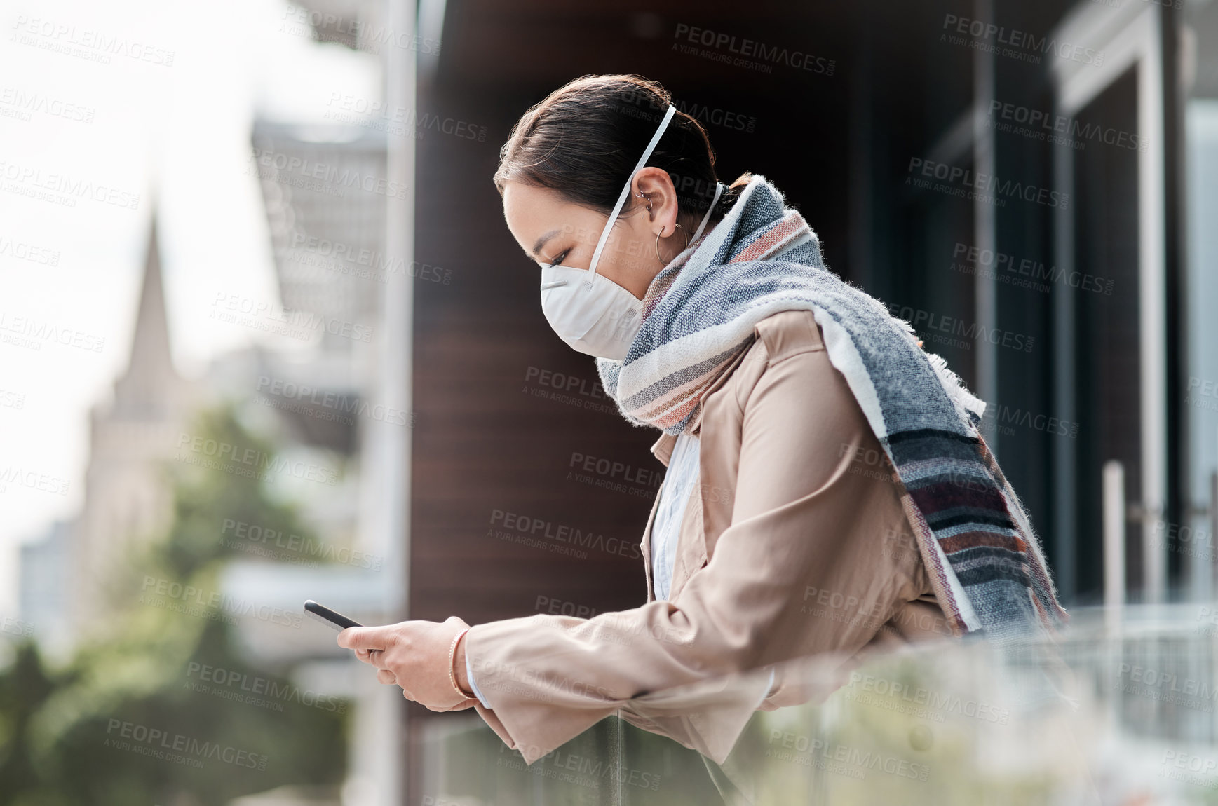 Buy stock photo Shot of a young woman wearing a mask and using a smartphone on the balcony at home