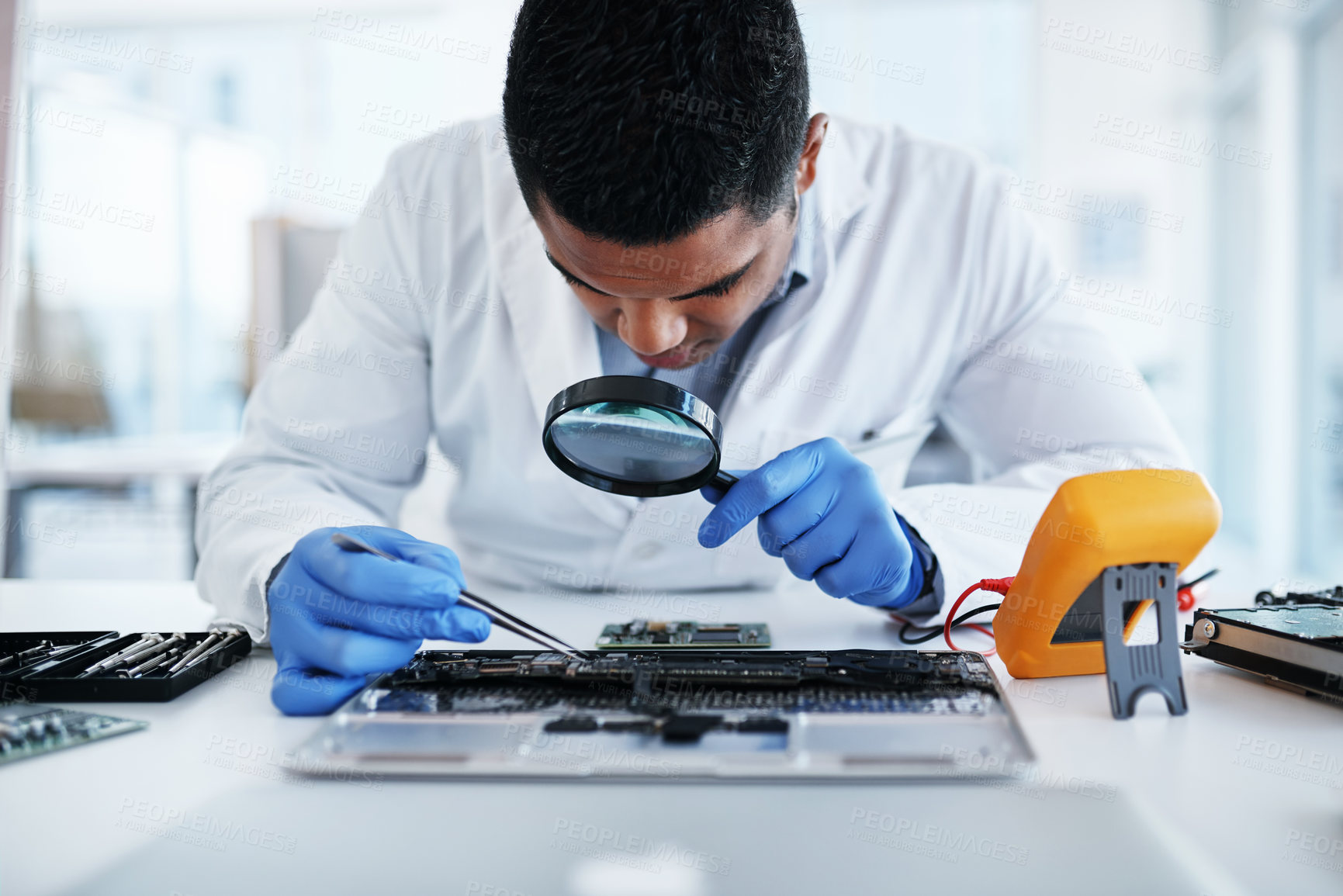 Buy stock photo Shot of a young man using tweezers and a magnifying glass while repairing computer hardware in a laboratory