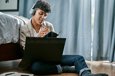 Buy stock photo Shot of a young man writing in a notebook and using a laptop while listening to music at home