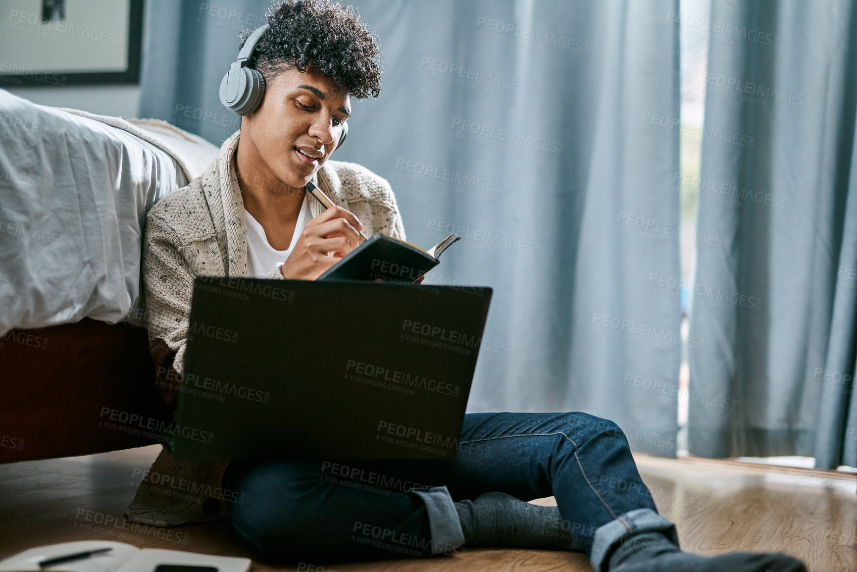 Buy stock photo Shot of a young man writing in a notebook and using a laptop while listening to music at home
