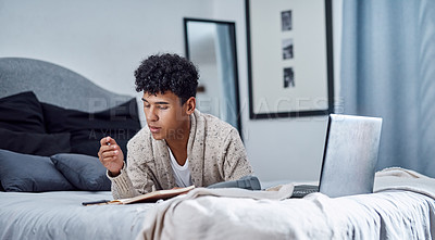 Buy stock photo Shot of a young man writing in a notebook and using a laptop on his bed at home