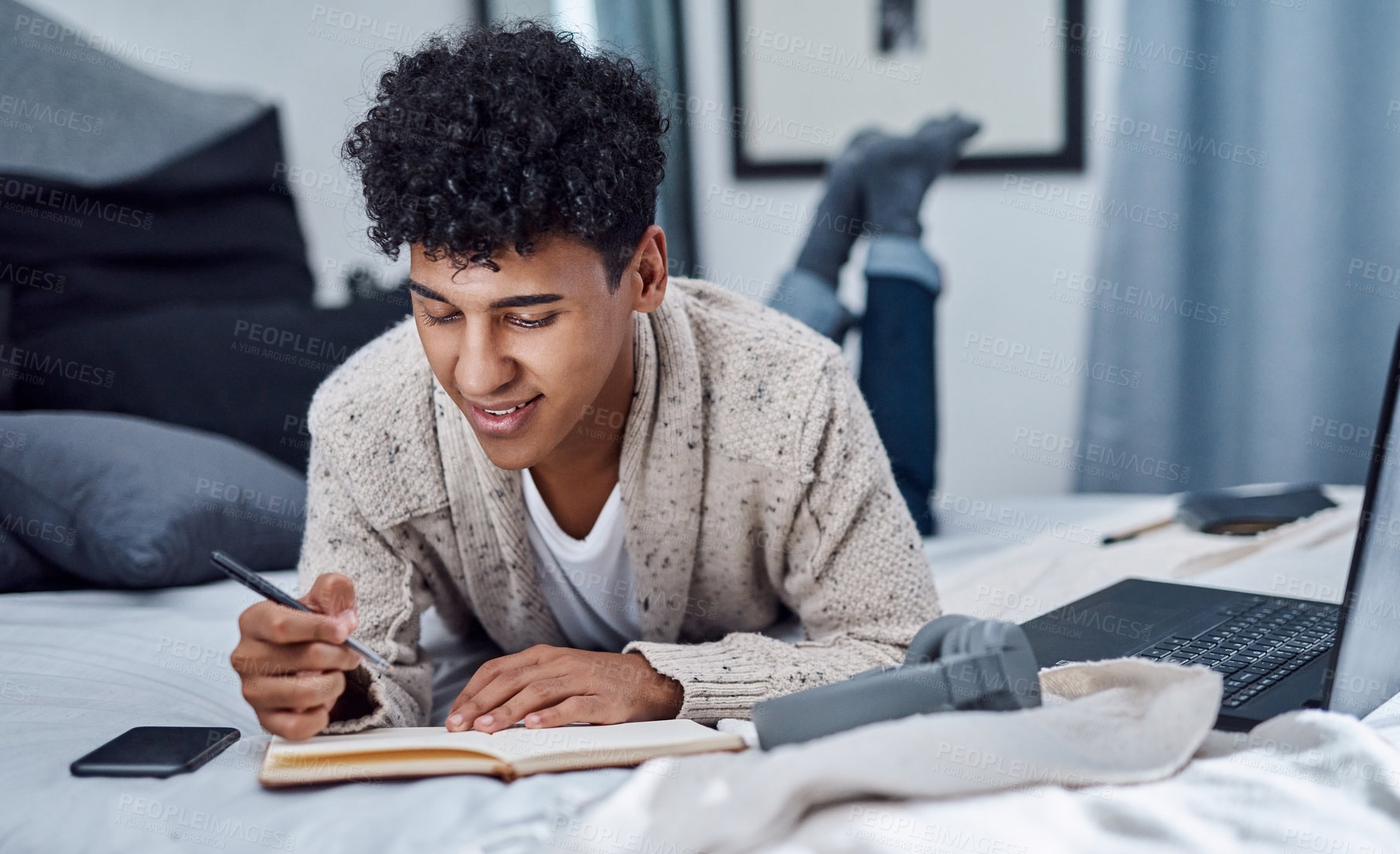 Buy stock photo Shot of a young man writing in a notebook and using a laptop on his bed at home