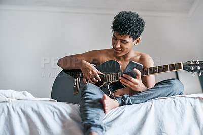 Buy stock photo Shot of a young man using a smartphone while playing the guitar at home