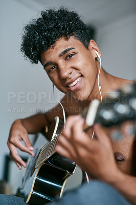 Buy stock photo Shot of a young man using earbuds while playing the guitar at home