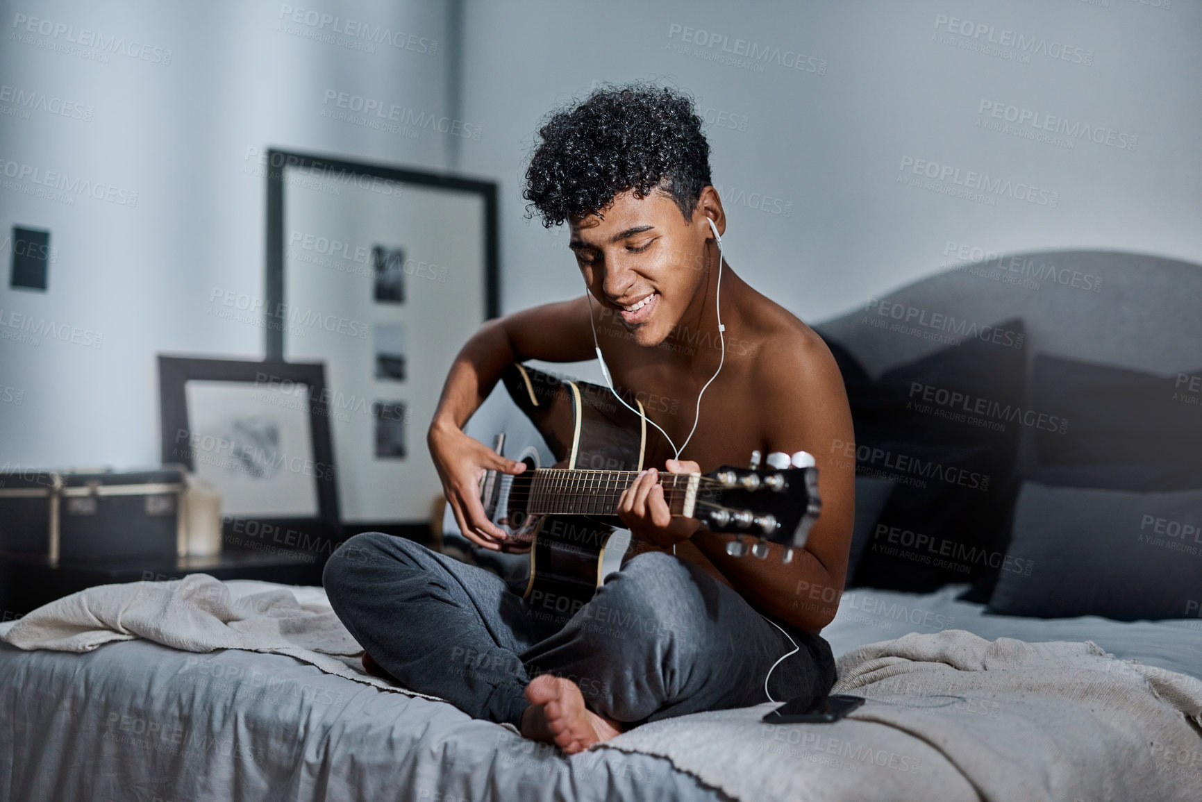 Buy stock photo Shot of a young man using a smartphone and earbuds while playing the guitar at home