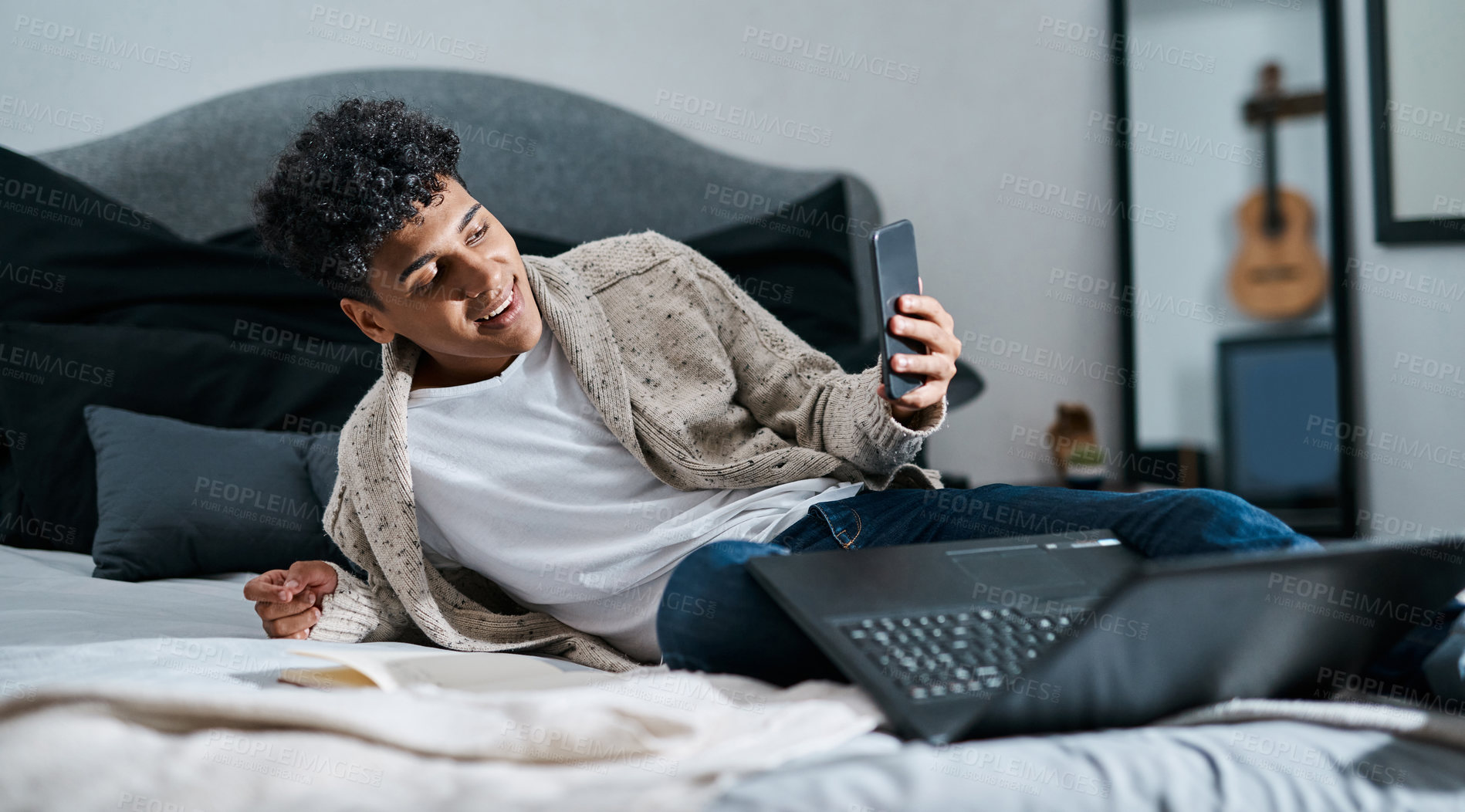 Buy stock photo Shot of a young man taking selfies using a smartphone while working with a laptop on his bed at home