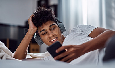 Buy stock photo Shot of a young man using a smartphone and headphones while relaxing on his bed at home