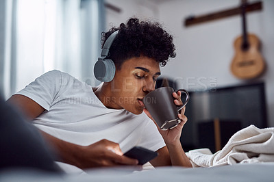 Buy stock photo Shot of a young man having coffee and using a smartphone with headphones while relaxing on his bed