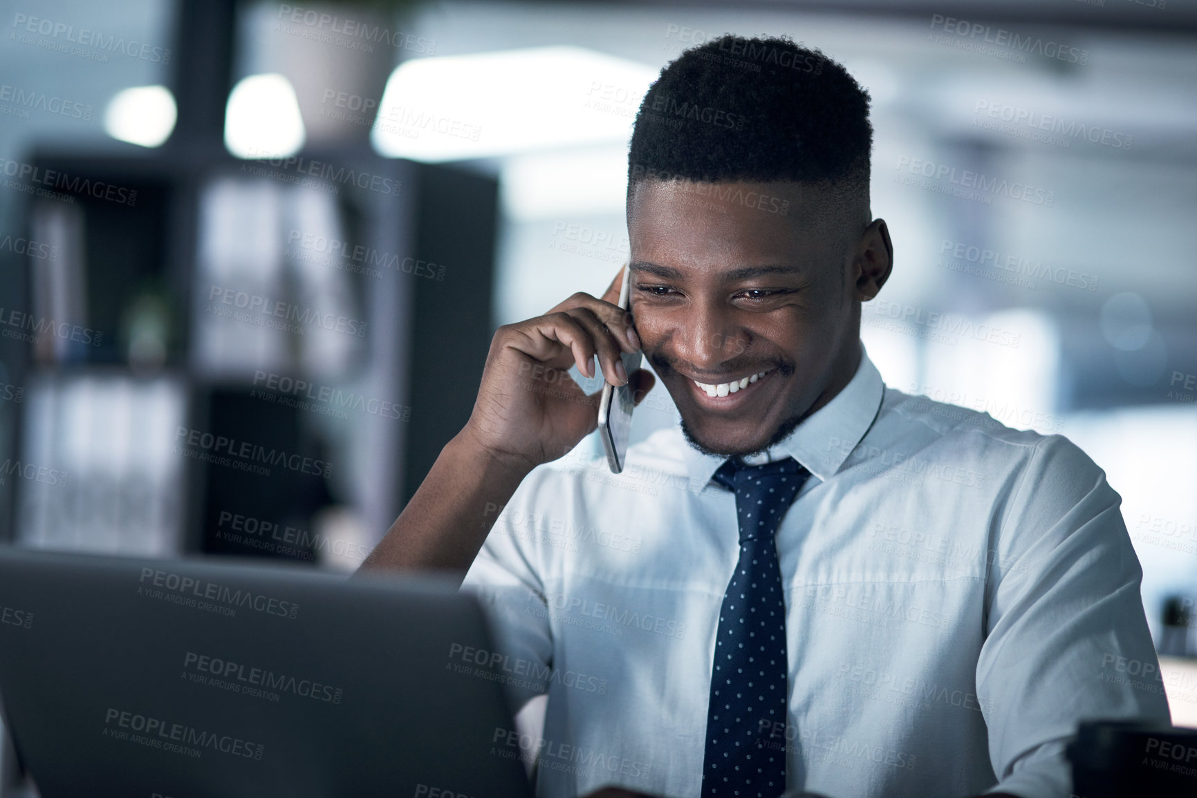 Buy stock photo Shot of a young businessman talking on a cellphone while working on a laptop in an office at night
