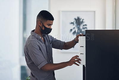 Buy stock photo Shot of a young businessman wearing a face mask while standing at a filling cabinet in an office