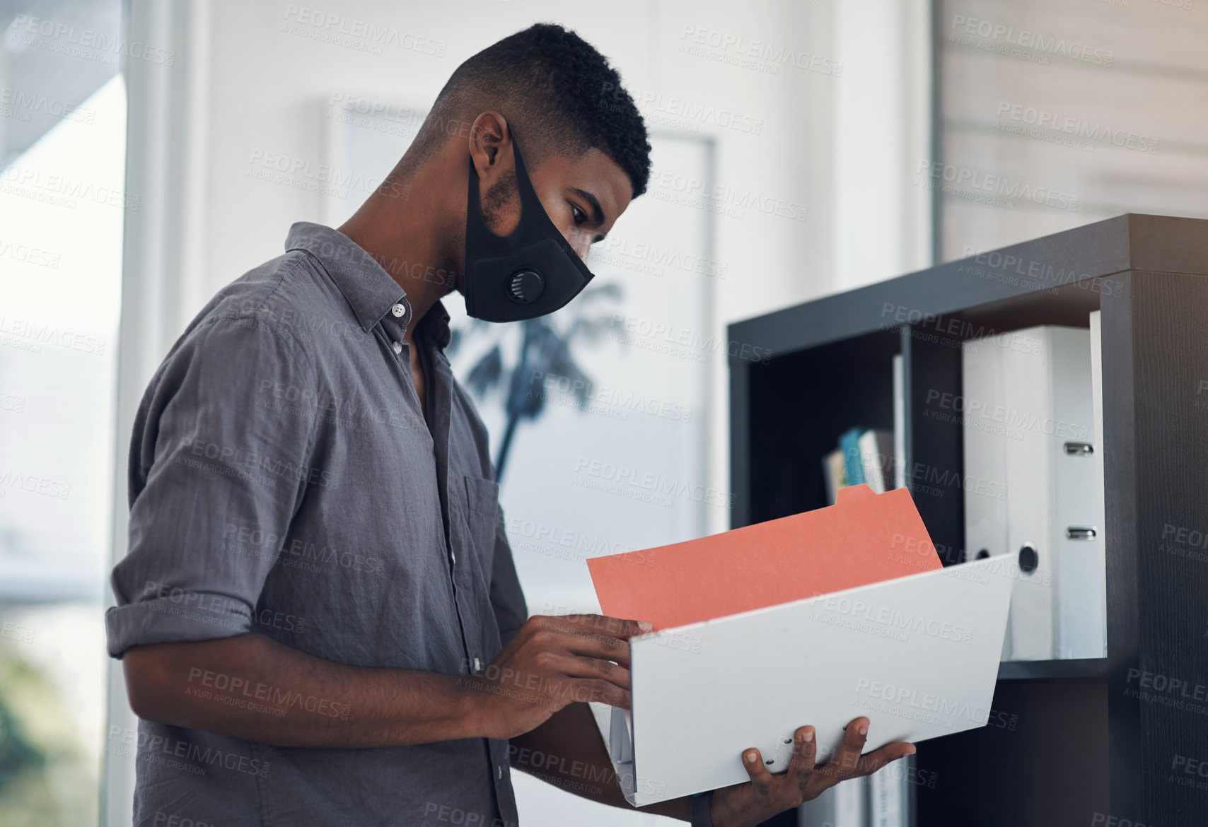 Buy stock photo Shot of a young businessman wearing a face mask while going through a folder in an office