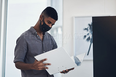 Buy stock photo Shot of a young businessman wearing a face mask while going through a folder in an office