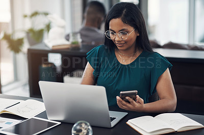 Buy stock photo Shot of a young designer using a laptop and cellphone in an office