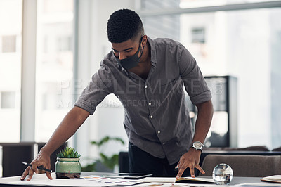 Buy stock photo Shot of a young designer wearing a face mask while going through paperwork in an office