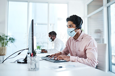 Buy stock photo Shot of a young call centre agent wearing a face mask while working on a computer in an office