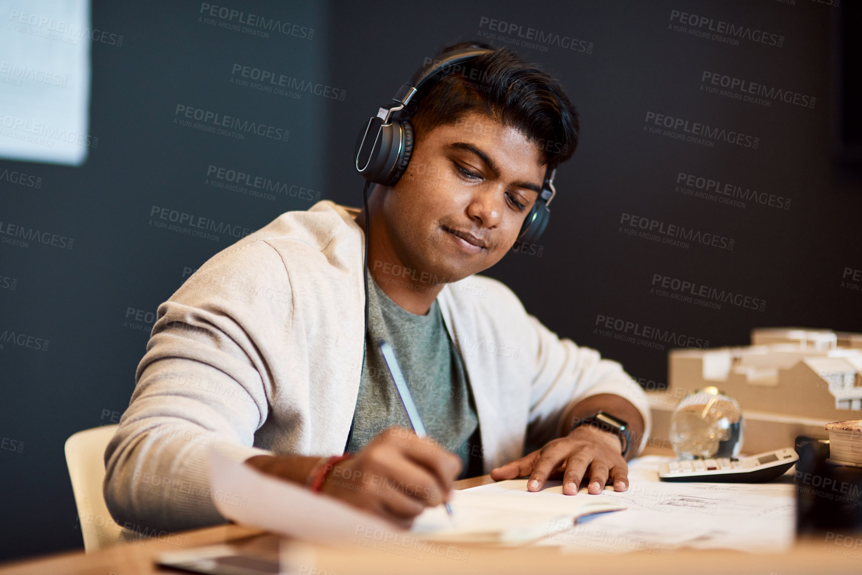 Buy stock photo Shot of a young architect wearing headphones and writing in a notebook at his desk in a modern office