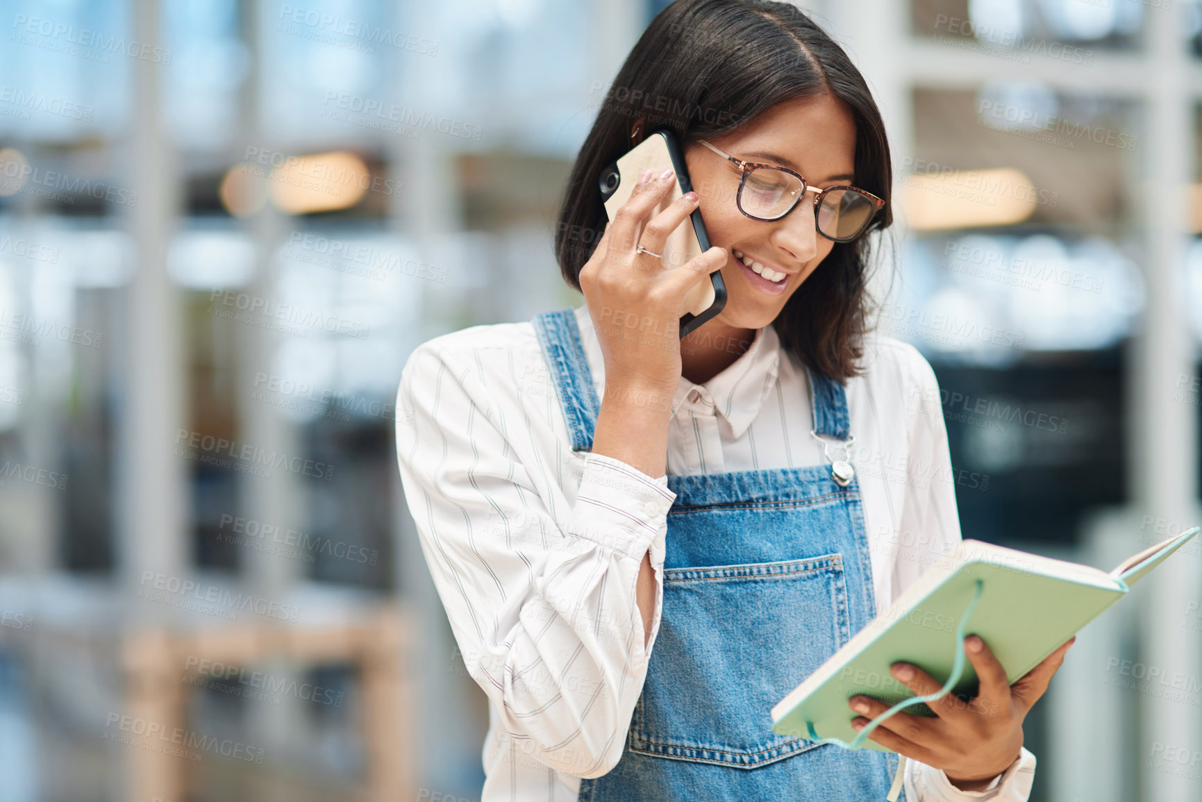 Buy stock photo Shot of a young businesswoman talking on a cellphone while going through a notebook in an office